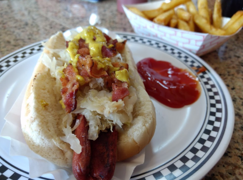 A hot dog topped with sauerkraut, bacon, and mustard, served with fries and ketchup on a checkered plate.