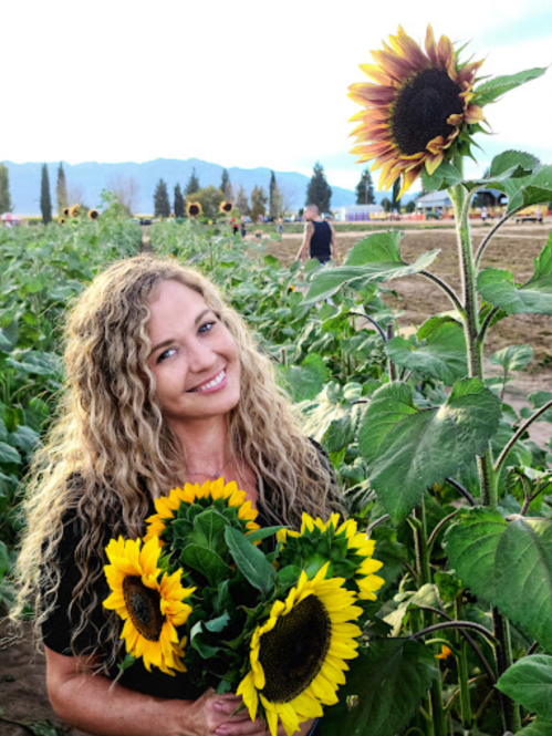 A woman with curly hair smiles while holding a bouquet of sunflowers in a field. Sunflowers and mountains are in the background.