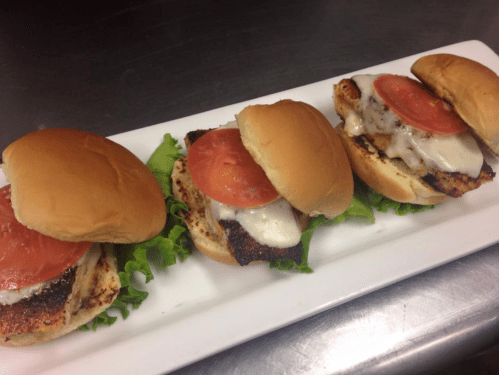 Three grilled chicken sandwiches with lettuce, tomato, and melted cheese on soft buns, served on a white platter.