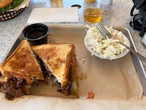 A toasted sandwich with meat and vegetables, served with a side of coleslaw and a small cup of sauce on a tray.