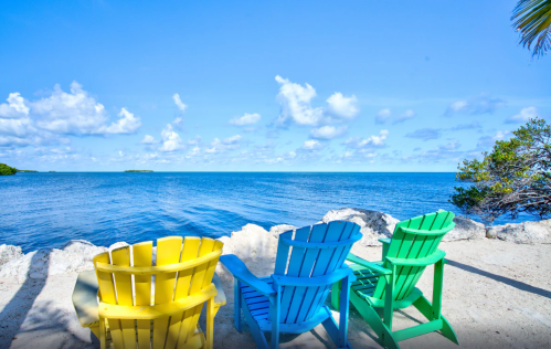 Three colorful Adirondack chairs face a calm blue ocean under a bright sky with fluffy clouds.