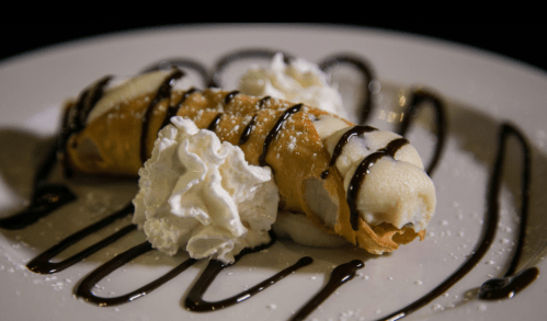 A delicious cannoli filled with cream, topped with whipped cream and drizzled with chocolate sauce on a white plate.