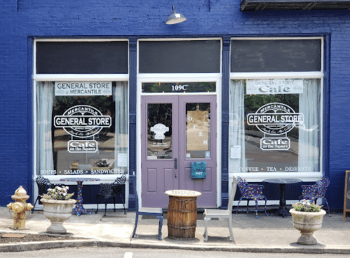 A quaint general store and café with purple doors, outdoor seating, and large windows displaying menu items.
