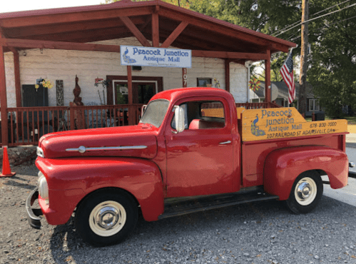 A vintage red pickup truck parked in front of the Peacock Junction Antique Mall, with a wooden sign on the back.