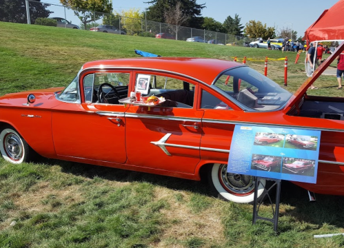 A vintage red car displayed at an outdoor event, with food on the window and an informational sign nearby.