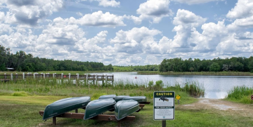 Canoes on a rack by a calm river, surrounded by lush greenery and a cloudy blue sky. Caution sign nearby.