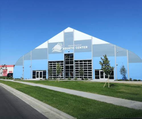 Exterior of the Idaho Central Aquatic Center, featuring a large, modern tent-like structure and green landscaping.