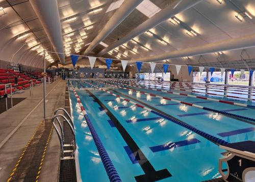 Indoor swimming pool with multiple lanes, blue and white flags, and seating area in a spacious, well-lit facility.