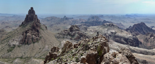 A panoramic view of rugged mountains and rocky terrain under a clear sky, with a person standing on a peak.
