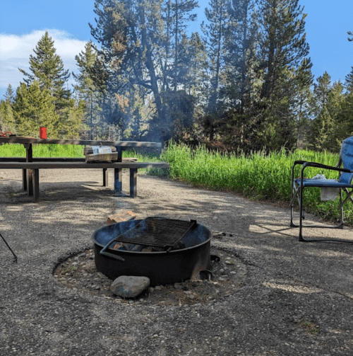 A campsite with a fire pit, grill, picnic table, and chairs surrounded by trees and green grass under a blue sky.