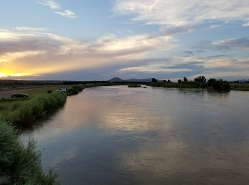 A serene river at sunset, reflecting colorful clouds, with lush greenery and distant mountains in the background.