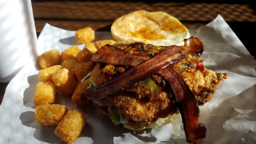 A crispy chicken sandwich topped with bacon, pickles, and lettuce, served with golden tater tots on the side.