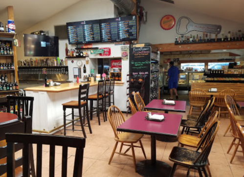 Interior of a casual dining space with wooden tables, chairs, a bar, and a menu board on the wall.