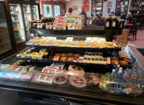 A display case filled with various cheeses, meats, and condiments in a deli, with customers seated in the background.