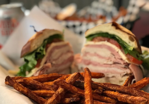 A close-up of a layered sandwich with lettuce, tomato, and meats, served with crispy sweet potato fries.