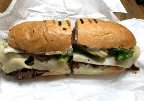 A freshly cut sandwich with roast beef, melted cheese, and lettuce on a toasted bun, resting on white paper.