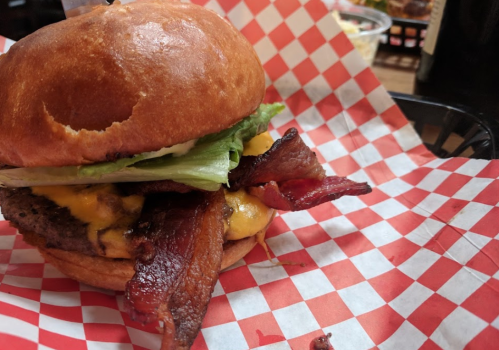 A close-up of a juicy burger with bacon, cheddar cheese, and lettuce on a toasted bun, served in a basket with red checkered paper.