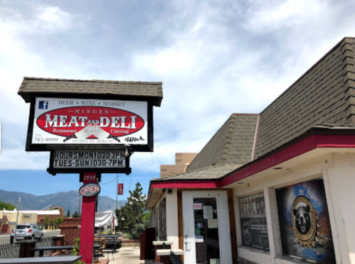 Sign for Minden Meat and Deli, featuring hours and an open sign, with a building in the background.