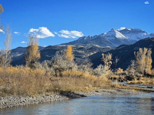 A serene river flows through a landscape of autumn trees and mountains under a clear blue sky.
