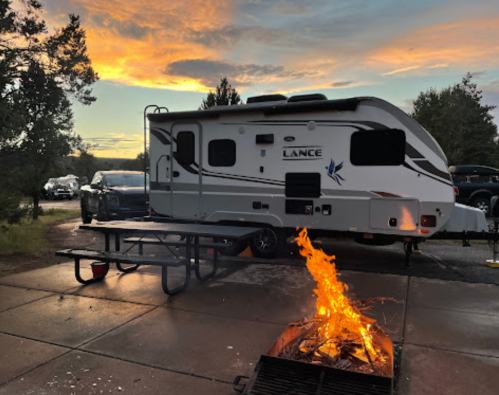 A campsite scene featuring a camper trailer, picnic table, and a fire pit with flames against a sunset sky.