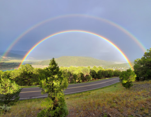 A vibrant double rainbow arcs over a green landscape and winding road under a cloudy sky.