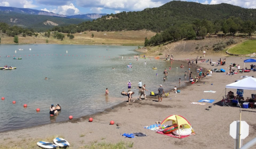 A sandy beach by a lake with people swimming, sunbathing, and enjoying various water activities under a clear sky.