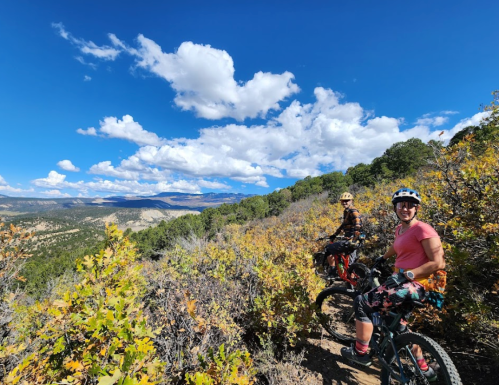 Two mountain bikers on a scenic trail surrounded by trees and vibrant autumn foliage under a blue sky with clouds.