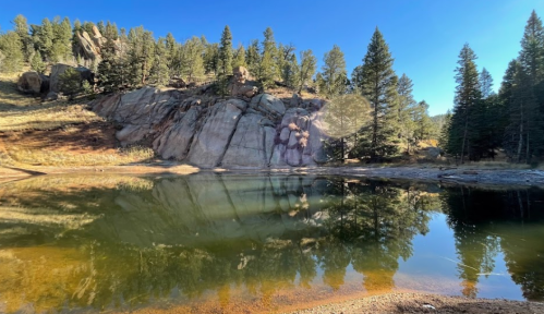 A serene pond reflecting trees and rocky terrain under a clear blue sky.