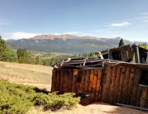 A weathered wooden structure in a mountainous landscape, with green trees and distant peaks under a clear blue sky.