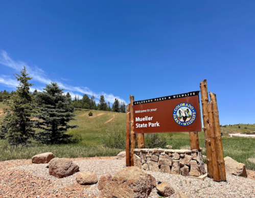 Sign welcoming visitors to Mueller State Park, surrounded by trees and a clear blue sky. Rocky terrain in the background.