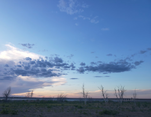 A serene landscape featuring a vast sky with clouds and a horizon at sunset, with bare trees in the foreground.