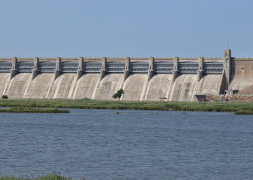 A large concrete dam stands against a clear blue sky, with water in the foreground and greenery along the shore.