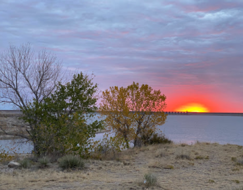 Sunset over a calm lake, with silhouetted trees and a colorful sky reflecting on the water's surface.