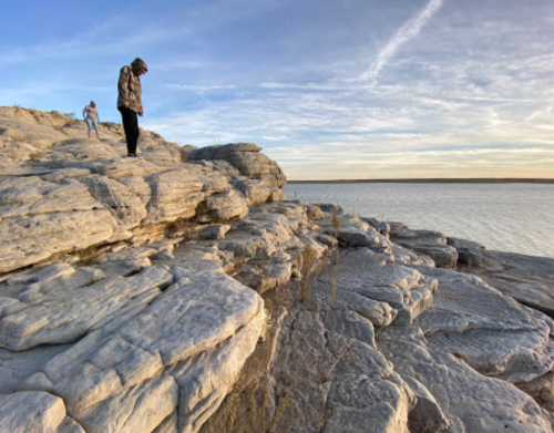 Two people explore rocky terrain by a calm body of water under a clear sky during sunset.