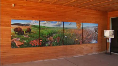 A mural depicting a vibrant landscape with bison, flowers, and a river, set against a wooden wall.
