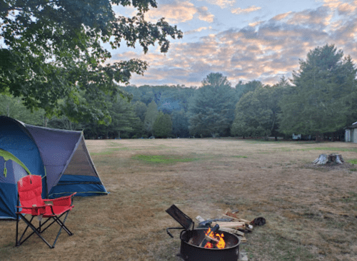 A campsite with a tent, a red chair, and a fire pit surrounded by trees and a cloudy sky.