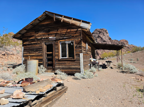 A rustic wooden cabin in a desert landscape, surrounded by rocks and sparse vegetation under a clear blue sky.