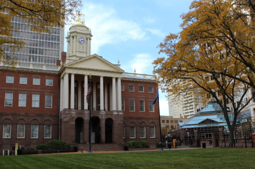 Historic building with a clock tower, surrounded by autumn trees and a green lawn, in an urban setting.