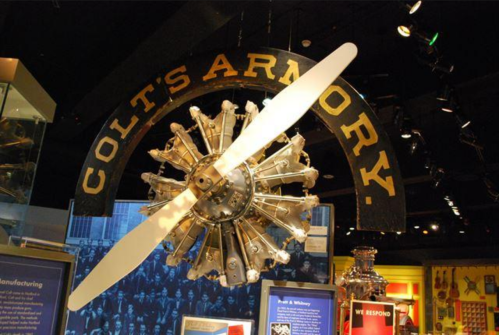 Sign reading "Colt's Armory" with a vintage aircraft propeller displayed in a museum setting.