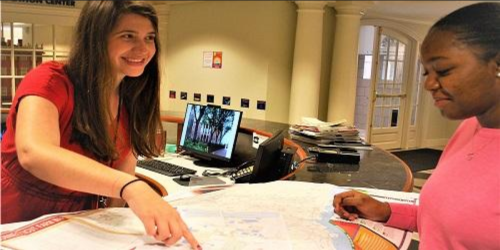 Two young women are discussing a map at a reception desk, with a computer and papers in the background.