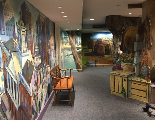Colorful murals decorate the walls of a hallway, featuring cityscapes and nature scenes, with a wooden bench in the foreground.