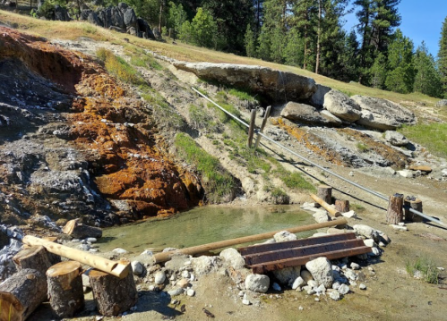 A rocky landscape with a small pond, wooden logs, and a metal cable, surrounded by trees and grass.