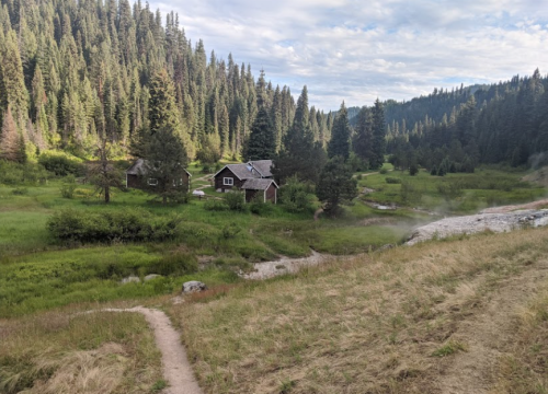 A serene landscape featuring a small cabin surrounded by lush greenery and tall trees, with a winding path and stream nearby.