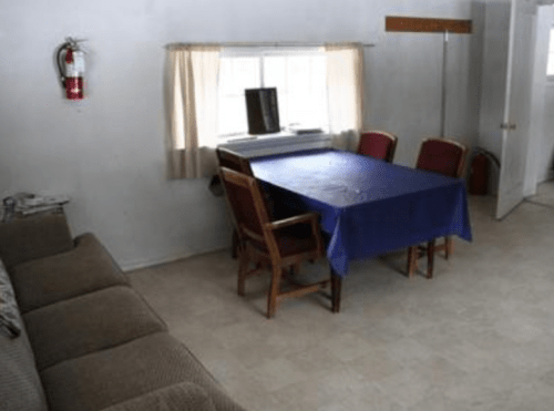 A simple room with a couch, a table covered with a blue cloth, and four chairs, featuring a window and a fire extinguisher.