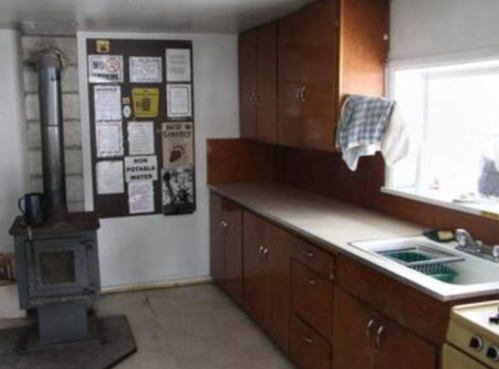 A small kitchen with wooden cabinets, a sink, and a wood stove, featuring a bulletin board on the wall.