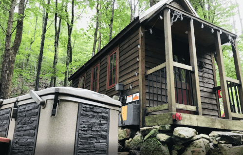 A small wooden cabin surrounded by trees, with a hot tub in the foreground on a rocky base.