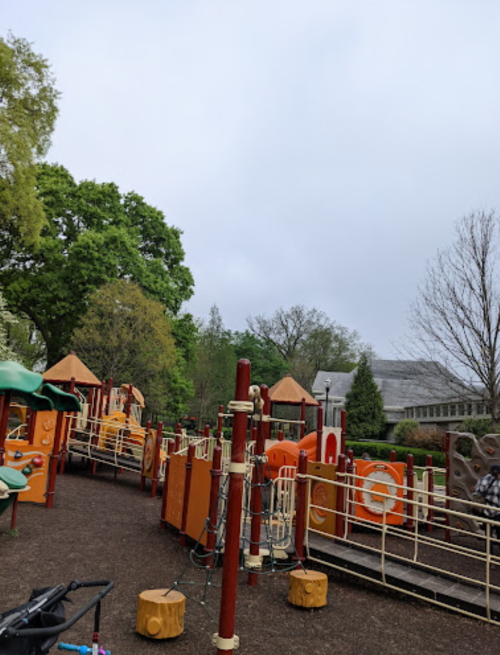 A playground with orange structures and wooden elements, surrounded by trees under a cloudy sky.