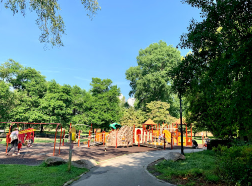 A sunny playground with colorful equipment surrounded by green trees and a walking path.