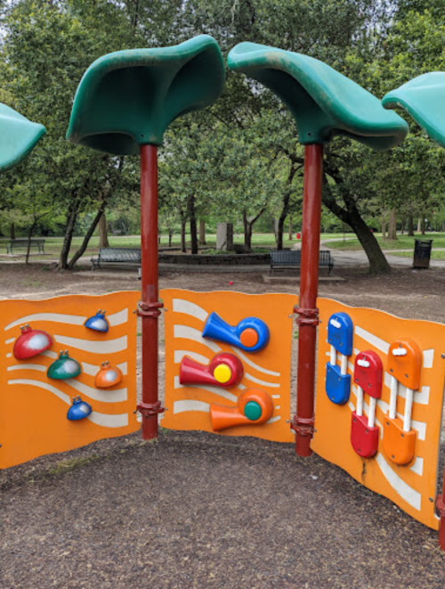 Colorful playground musical instruments mounted on a wall, surrounded by green trees in a park setting.