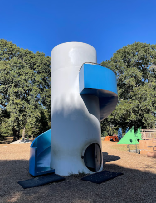 A cylindrical playground structure with a blue slide and a small entrance, surrounded by trees and play equipment.